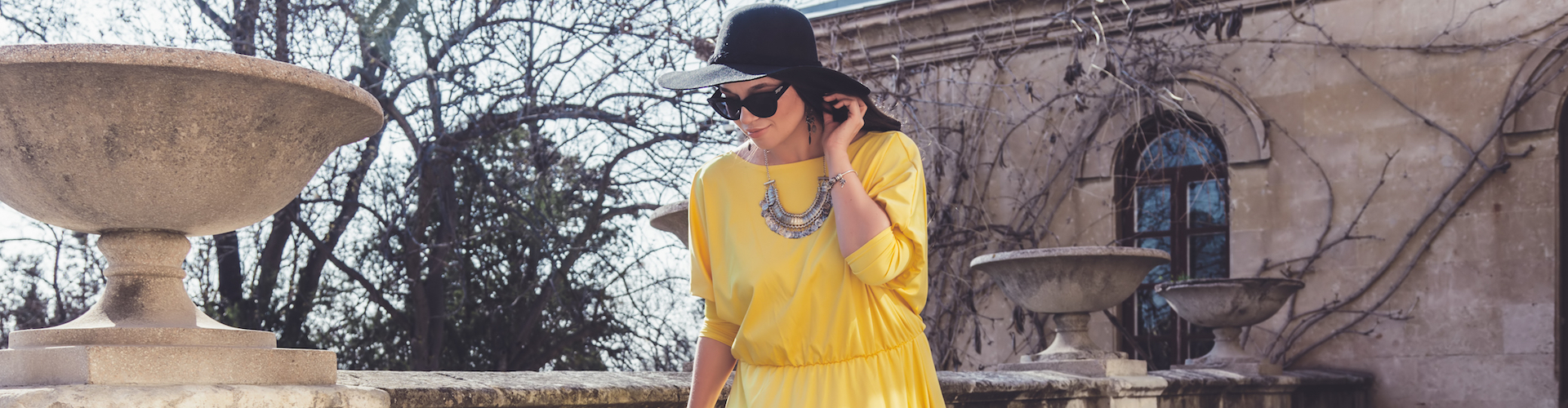 Young stylish woman wearing yellow maxi dress, black hat and sunglasses walking in the city street